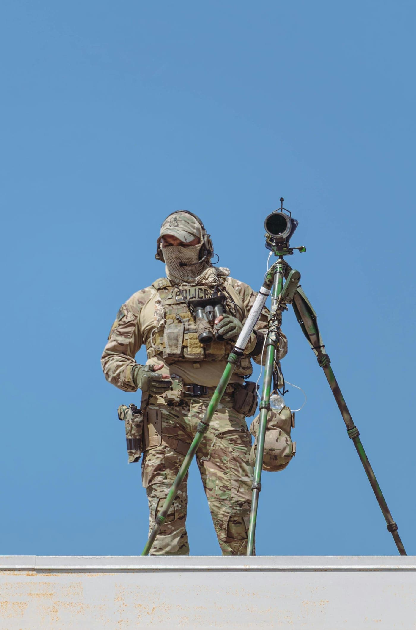Law-enforcement officer in tactical uniform standing beside a tripod-mounted observation scope against a clear blue sky.